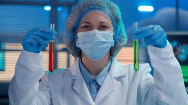 Bir kadın bilim adamı elinde kırmızı ve yeşil sıvıyla test tüpleri tutuyor. Mavi eldivenli bir kadın portresi, beyaz bir tıbbi elbise, şapka ve maske. Modern bir biyokimya laboratuvarında araştırma