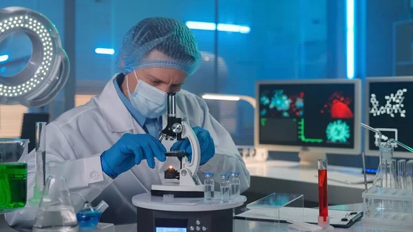 一名身穿白袍 戴着面具 戴着蓝色手套和帽子的男性研究人员正在显微镜下对样本进行检测 穿着生化工作服的人都在后台工作 研究生化的工作流程 — 图库照片
