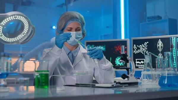 一位女科学家拿着长笛 把绿色液体吸入试管 女医生或研究人员穿着白袍 蓝色手套和帽子 带有试管的现代生化实验室 — 图库照片