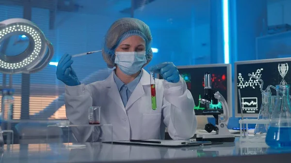 一位女科学家用绿色液体从管道里把一滴红色的物质扔进试管里 身穿白袍 戴着面具 戴着蓝色手套和帽子的女人观察了样本的反应 — 图库照片