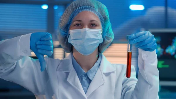 A woman scientist holds a test tube with a red liquid in her hand and shows a thumbs down. A woman in blue gloves, a white medical gown and a mask shows a dislike gesture, bad, dangerous. Close up