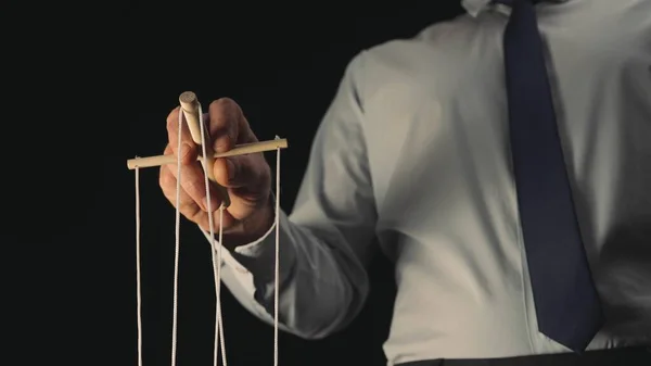 A businessman in a gray shirt and black tie controls a puppet with a wooden manipulator and strings. A puppeteer controls a doll marionette on a black isolated background. The concept of control