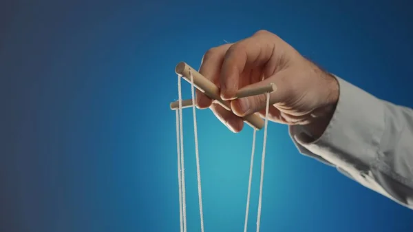 The hand of a man in a gray shirt controls a puppet using a wooden manipulator and strings on a blue background. Hand handling at puppet by pulling strings to make the character move. The concept of