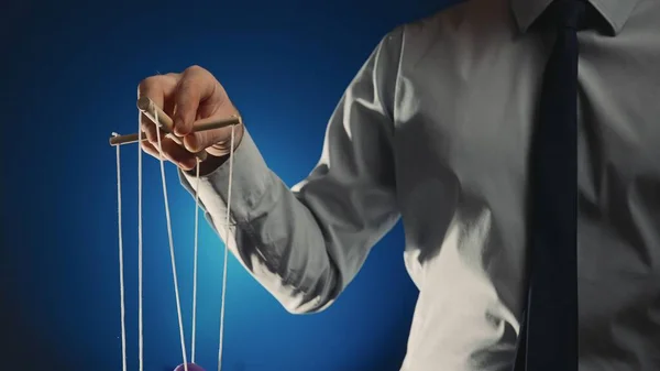 A businessman in a gray shirt and black tie controls a puppet with a wooden manipulator and strings. Hand handling at puppet by pulling strings to make the character move. Blue isolated background