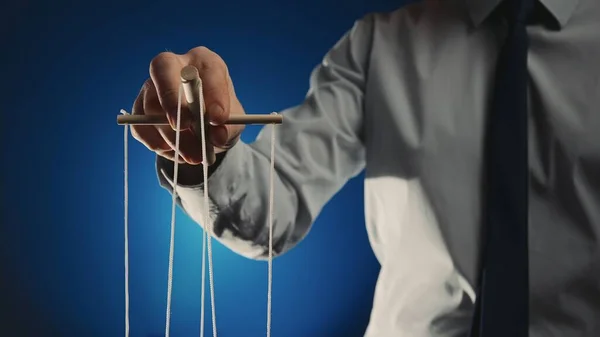 A businessman in a gray shirt and black tie controls a puppet with a wooden manipulator and strings. Hand handling at puppet by pulling strings to make the character move. Blue isolated background