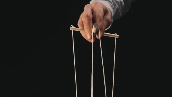 The puppeteers hand controls the puppet with a wooden manipulator and strings. The marionettist controls and pulls the strings on a black background. The concept of dependence, dominance, managing