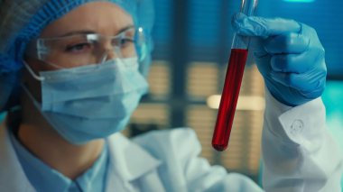 A woman carefully examines the red liquid in a test tube. Female doctor or researcher in goggles, bonnet, mask, white gown and blue gloves examines a sample. Biochemical laboratory, hospital. The