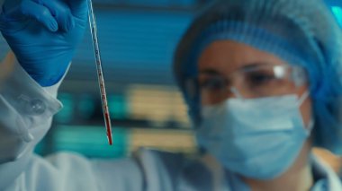 Bir kadın kırmızı sıvıyla dolu cam bir pipeti dikkatle inceliyor. Gözlüklü, boneli, maskeli, beyaz önlüklü ve mavi eldivenli kadın doktor veya araştırmacı biyokimya laboratuvarında veya hastanede. Mavi ışık