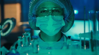 Tıbbi maskeli, gözlüklü, beyaz önlüklü ve mavi şapkalı bir kadın doktor portresi. Bir kadın cam mataralar, test tüpleri ve şişelerden doğrudan kameraya bakar. Modern laboratuvar ya da