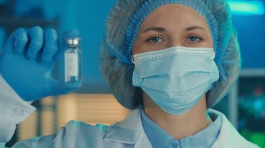Beyaz elbiseli, tıbbi maskeli, boneli ve mavi eldivenli bir kadının portresi elinde aşı ya da ilaç içeren cam bir şişe tutuyor. Kadın bir doktor arka plandaki kameraya bakar.