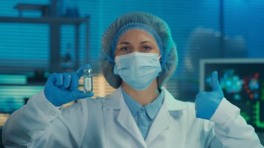 Bir kadın elinde cam aşı şişesi tutuyor ve baş parmağını kaldırıyor. Bir laboratuvar ya da hastanenin arka planında beyaz önlüklü, tıbbi maskeli, boneli ve mavi eldivenli bir kadın doktor portresi.