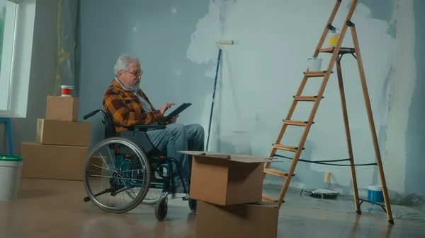 An elderly disabled man moves in a wheelchair plans renovation using a digital tablet. Room with window, ladder, cardboard boxes, wallpaper rolls, paint bucket. Concept of repair in the apartment.