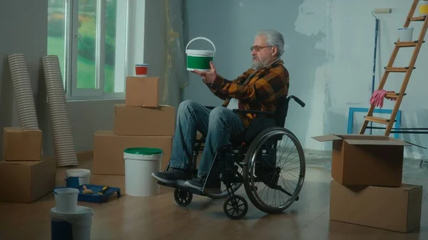 一个坐在轮椅上的老年残疾人正在检查一桶油漆 养恤金领取者计划进行修理和墙壁装饰 有窗户 壁纸卷的房间 联合国系统内的修理概念 — 图库照片