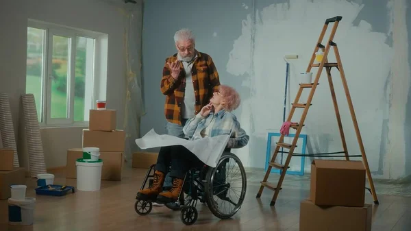 一位坐在轮椅上的老年男女正在看一张有公寓项目的单子 一对年迈的夫妇正计划在房子里靠窗 纸板进行维修 — 图库照片