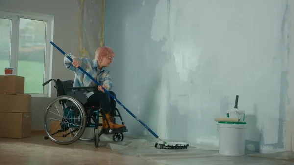 一位坐在轮椅上的老年妇女用长长的滚筒把墙壁涂上了白漆 一位年事已高的残疾妇女在房间里修理 人们在公寓里进行维修的概念 — 图库照片