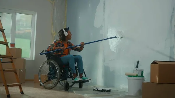 An African American woman in white headphones on a wheelchair paints a wall with white paint using a long roller. A young black woman with a disability makes repairs in room and enjoys music. The
