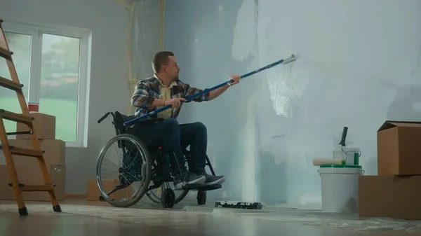 A young man in a wheelchair paints a wall with white paint using a long roller. A disabled man makes repairs in the room against the background of cardboard boxes and buckets of paint. The concept of