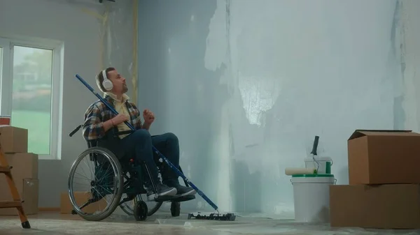 A young man in white headphones on a wheelchair plans to paint a wall with white paint using a long roller. A disabled man makes repairs in the room and enjoys music. The concept of renovation in the