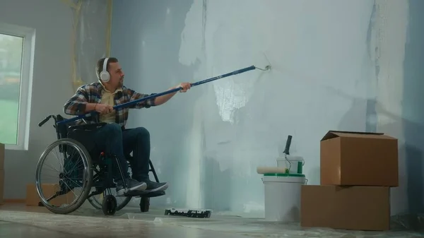 A young man in white headphones on a wheelchair paints a wall with white paint using a long roller. A disabled man makes repairs in the room and enjoys music. The concept of renovation in the