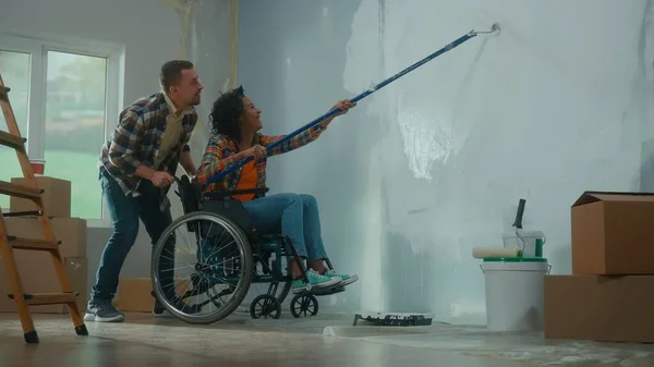 An African American woman in a wheelchair paints a wall with white paint using a long roller. A black woman and a white man make repairs in the room and laugh happily. A young couple. The concept of