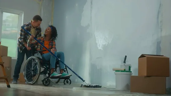 An African American woman in a wheelchair paints a wall with white paint using a long roller. A black woman and a white man make repairs in the room and laugh happily. A young couple. The concept of