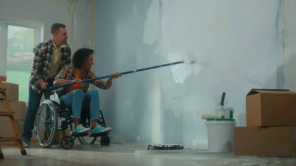 一位坐在轮椅上的非洲裔美国妇女用一个长滚筒画了一堵白漆墙 一个黑人女人和一个白人男人在房间里修理 开心地笑着 一对年轻夫妇一 — 图库照片