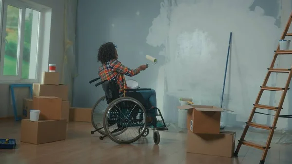 An African American woman on a wheelchair planning to paint the wall with paint using a roller. View from the back of the young black woman with a disability makes repairs in room. The concept of