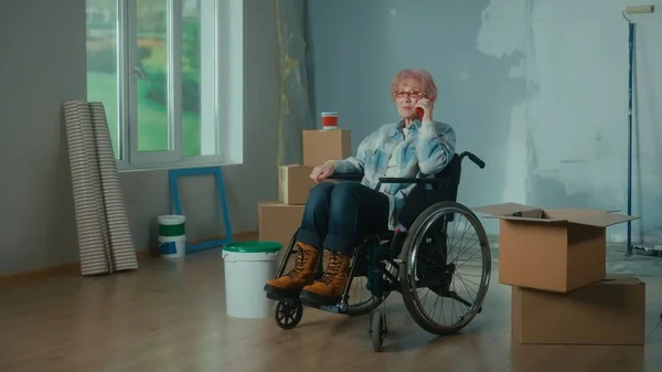 一位坐在轮椅上的老年残疾妇女用手机交谈 已退休的残疾女性 有窗户的房间 油漆桶 壁纸卷 翻修的概念 — 图库照片