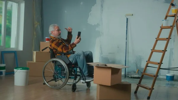 An elderly disabled man in a wheelchair talks on a video call using a mobile phone. A pensioner male points to the window. A room with window, wallpaper rolls, cardboard boxes and a bucket of paint
