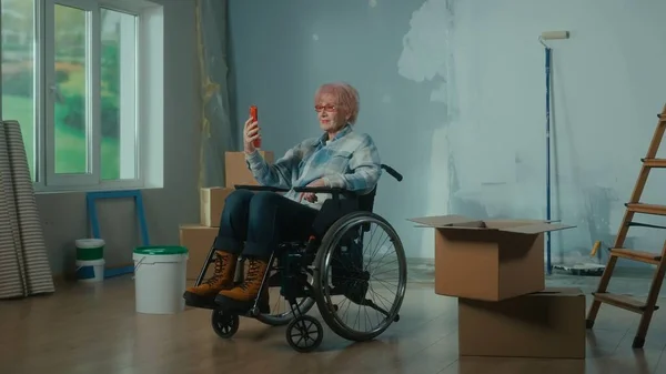 An elderly disabled woman in a wheelchair talks on a video call using a mobile phone. A room with a window, ladder, wallpaper, cardboard boxes and a bucket of paint. Concept of repair in the apartment