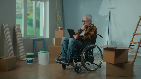 An elderly disabled man moves in a wheelchair plans renovation using a digital tablet. Room with window, ladder, cardboard boxes, wallpaper rolls, paint bucket. Concept of repair in the apartment.