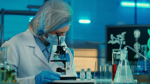 A female scientist examines a sample under a microscope. A woman doctor in a white medical gown, bonnet, blue gloves and a mask. Laboratory with test tubes, flasks and computer monitors in blue light