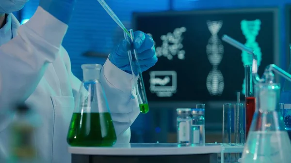 女性科学者がガラス試験管に緑色の液体をピペットします 白い医療用のガウンを着た女性 ボンネット 青い手袋と青色で試験管 フラスコやコンピュータモニタを備えた研究室のマスク — ストック写真