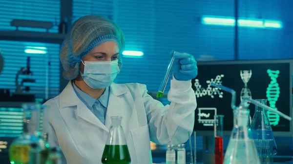 一位女科学家正在对玻璃管中的绿色液体进行审查和研究 一名身穿白色医疗服 戴着帽子 戴着蓝色手套 戴着面具 戴着试管 瓶子和口罩的妇女 — 图库照片