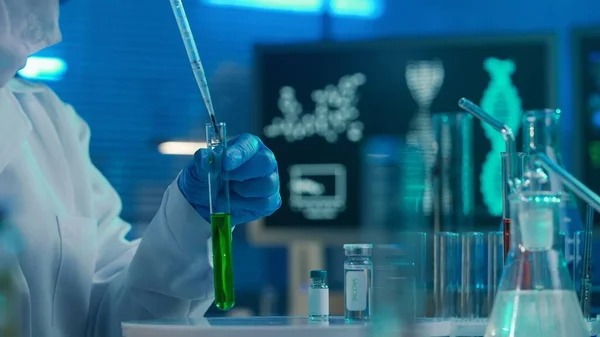 一位女科学家把绿色液体塞入玻璃试管 一名身穿白衣 戴着帽子 戴着蓝色手套 戴着面具 戴着试管 瓶子和蓝色计算机显示器的妇女 — 图库照片