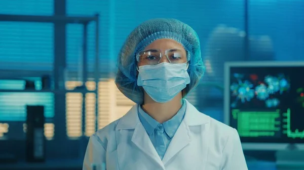 Portrait of a woman doctor in a medical mask, goggles, white gown and blue bonnet looks directly into the camera. Female scientist or researcher posing against the backdrop of a modern laboratory or