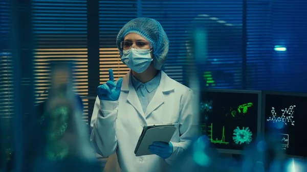 女医生检查玻璃瓶 并将数据输入数字平板电脑 穿着白色长袍 护目镜 面罩和手套的女科学家 在实验室或医院里 带着蓝光 — 图库照片