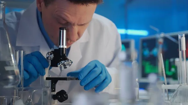 一名身穿白色长袍和蓝色手套的男性研究人员正在显微镜下检查样本 有许多试管和玻璃瓶的实验室或医院 在后台是一个计算机监视器 工作流程 — 图库照片