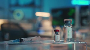 Ön planda, içinde tıbbi aşı ve şırınga bulunan cam şişeler var. DNA yapısı ve virüsler bilgisayar ekranlarında görülebilir. Laboratuvar asistanı ya da araştırmacısının boş işyeri. Modern