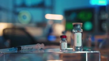 Ön planda, içinde tıbbi aşı ve şırınga bulunan cam şişeler var. DNA yapısı ve virüsler bilgisayar ekranlarında görülebilir. Laboratuvar asistanı ya da araştırmacısının boş işyeri. Modern