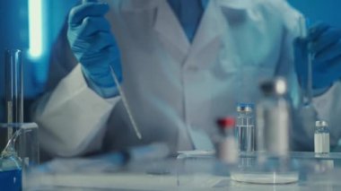 Ön planda aşı ve şırıngayla birlikte cam şişeler var. Beyaz elbiseli, tıbbi maskeli ve mavi eldivenli bir erkek doktor cam pipetten bir damla yeşil sıvıyı test tüpüne damlatıyor. Şey...