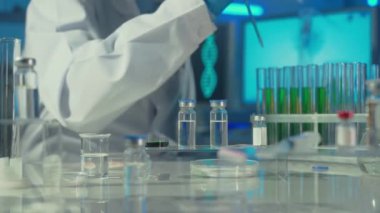 Laboratuvar araştırmacısı cam pipetten bir damla yeşil maddeyi sıvı ile test tüpüne damlatıyor. Araştırma verilerini beyaz önlüklü ve mavi eldivenli bir erkek doktor yazıyor. Aşılı cam şişeler ve...