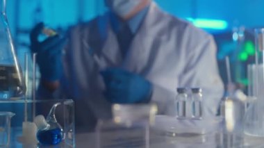 Ön planda COVID 19 tıbbi aşı tüpleri odaklanmış durumda. Beyaz önlüklü, tıbbi maskeli, gözlüklü ve mavi eldivenli bir adam aşıyı şırıngaya çeker. Modern biyokimyasal araştırma