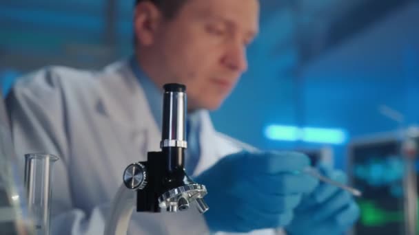 男性科学者はガラスのピペットでサンプルを適用し 顕微鏡下で検査します 研究者はその結果をコンピュータに入力する 前景では 医療用ワクチンボトルが注目されています — ストック動画