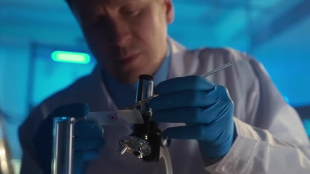 男性科学者はガラスピペットで液体または血液の赤いサンプルを適用し 顕微鏡下でそれを検査します 前景では Covid 19医療用ワクチンのバイアルが注目されている — ストック動画