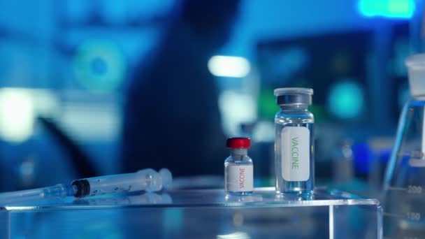 前景光明的是装有医疗疫苗和注射器的玻璃瓶 一名身穿白色长袍 戴着医疗面具 戴着护目镜 戴着蓝色手套的男医生正在电脑键盘上打字 现代研究 — 图库视频影像