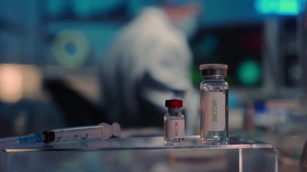 前景光明的是装有医疗疫苗和注射器的玻璃瓶 一名身穿白色长袍 戴着医疗面具 戴着护目镜 戴着蓝色手套的男医生正在电脑键盘上打字 现代研究 — 图库视频影像