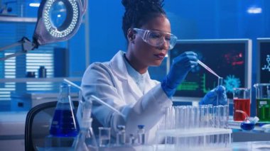 Plastik pipet kullanan genç bir Afrikalı Amerikalı kadın kırmızı ve yeşil sıvıyı cam test tüplerine koyuyor ve numuneleri inceliyor. Siyah bir kadın doktor beyaz önlüklü, gözlüklü ve eldivenli.