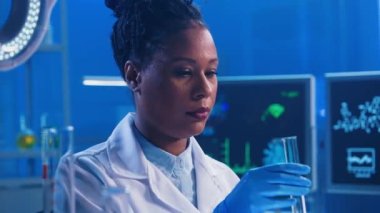 Genç bir Afrikalı Amerikalı kadın yeşil bir sıvıyı plastik bir pipetle bir şişeye dolduruyor, kameraya bakıyor ve gülümsüyor. Beyaz önlüklü ve eldivenli siyahi bir kadın doktor modern bir yerde çalışıyor.