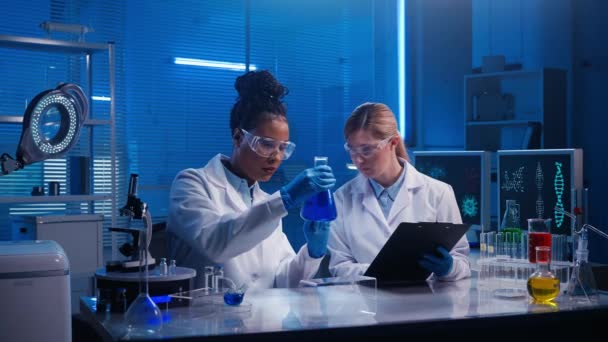 一位非洲裔美国女医生检查了瓶中的蓝色液体 而一位白人女性实验室助理写下了数据 身穿白衣 蓝色手套和护目镜的医护人员在实验室工作 — 图库视频影像